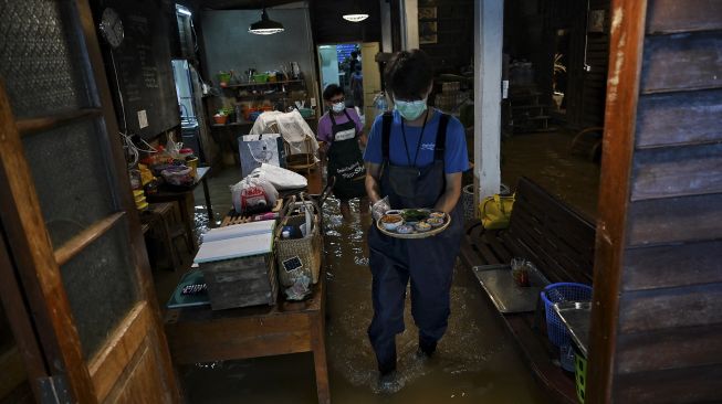 Seorang karyawan yang menyajikan makanan di Kafe Antik Chaopraya, saat air banjir dari Sungai Chao Phraya mengalir ke restoran di provinsi Nonthaburi, Bangkok, Thailand, pada (7/10/2021). [Lillian SUWANRUMPHA / AFP]