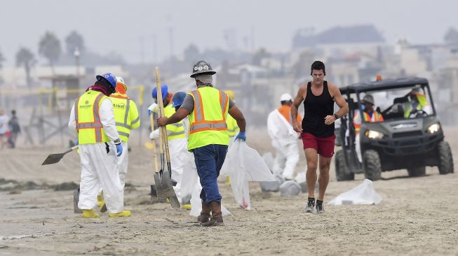 Seorang pengunjung berlari melewati kru pembersihan yang bekerja di Pantai Newport, California, pada (7/10/2021). [Frederic J. BROWN / AFP]