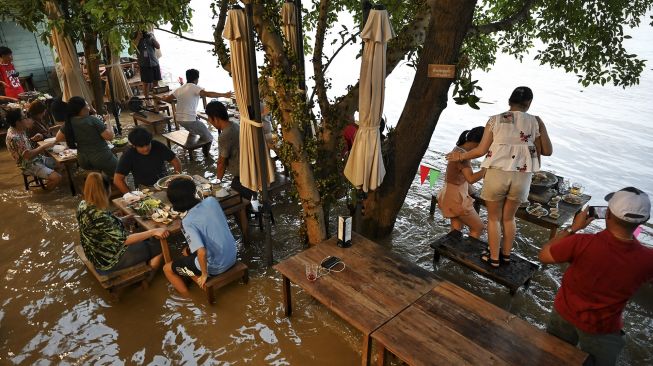 Pengunjung berdiri sambil menikmati makan malam di Kafe Antik Chaopraya, saat air banjir dari Sungai Chao Phraya mengalir ke restoran di provinsi Nonthaburi, Bangkok, Thailand, pada (7/10/2021). [Lillian SUWANRUMPHA / AFP]