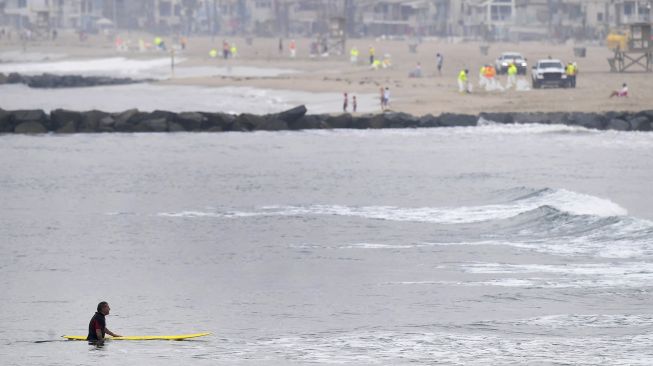 Seorang peselancar berdiri di air dengan latar belakang kru pembersihan yang terus bekerja setelah tumpahan minyak di Pantai Newport, California, pada (7/10/2021). [Frederic J. BROWN / AFP]