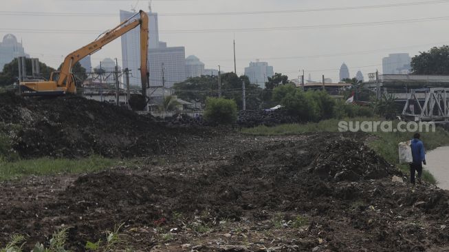 Warga berjalan di dekat alat berat yang melakukan pengerukan sedimen lumpur di sepanjang aliran Kanal Banjir Barat di kawasan Tanah Abang, Jakarta, Jumat (29/10/2021). [Suara.com/Angga Budhiyanto]