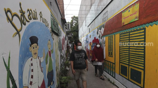 Warga berjalan melintasi mural bertema budaya Betawi di kawasan Kampung Sentra Kreatif, RW 006 Kota Bambu Selatan, Palmerah, Jakarta, Rabu (3/11/2021). [Suara.com/Angga Budhiyanto]