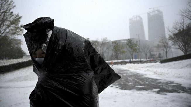 Seorang warga menggunakan kantong plastik sebagai penutup saat salju turun di Beijing, China, Minggu (7/11/2021). [NOEL CELIS / AFP]