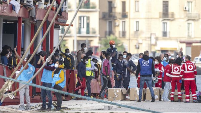 Relawan dari Palang Merah Italia (kanan), Organisasi Internasional untuk Migrasi (IOM) dan UNICEF (kiri) menyambut para migran yang diselamatkan di Laut Mediterania menggunakan kapal Sea-Eye 4 setibanya di Pelabuhan Trapani, Sisilia, Italia, pada (8/11/2021). [GIOVANNI ISOLINO / AFP]