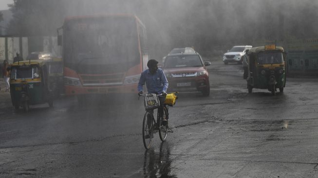 Para komuter melintas melewati alat anti-kabut asap yang menyemprotkan air untuk mengekang polusi udara di New Delhi, India, pada (16/11/2021). [MONEY SHARMA / AFP]