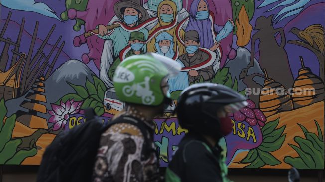 Pengendara sepeda motor melintas di depan mural bertema COVID-19 di Jakarta, Jumat (26/11/2021). [Suara.com/Angga Budhiyanto]