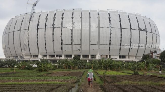 Seorang petani berjalan di area lahan pertanian dengan latar belakang proyek pembangunan Jakarta International Stadium (JIS) di kawasan Sunter Agung, Jakarta, Kamis (23/12/2021). [Suara.com/Angga Budhiyanto]