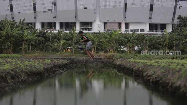 Seorang anak memancing ikan di area lahan pertanian dengan latar belakang proyek pembangunan Jakarta International Stadium (JIS) di kawasan Sunter Agung, Jakarta, Kamis (23/12/2021). [Suara.com/Angga Budhiyanto]