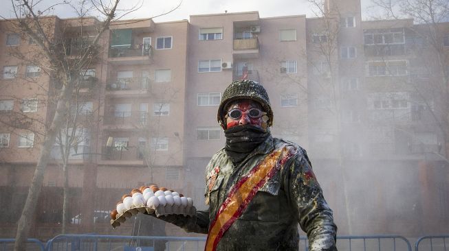 Orang yang bersuka ria mengenakan pakaian militer memegang telur saat pertempuran "Els Enfarinats" di Kota Ibi, Spanyol, pada (28/12/2021). [JAIME REINA / AFP]