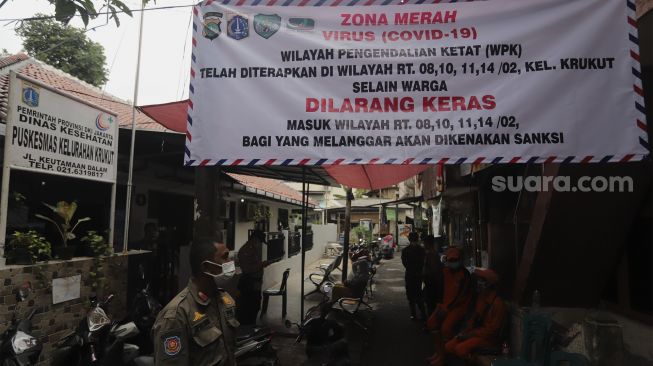 Petugas berdiri di dekat spanduk tanda zona merah COVID-19 di RW 02 Kelurahan Krukut, Kecamatan Taman Sari, Jakarta, Senin (10/1/2021). Wilayah tersebut kekinian diterapkan micro lockdown. [Suara.com/Angga Budhiyanto]