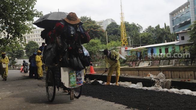 Pedagang keliling melintas di samping perbaikan jalan ambles di Jalan Inspeksi Kali Ciliwung, Kenari, Kecamatan Senen, Jakarta, Rabu (19/1/2022). [Suara.com/Angga Budhiyanto]