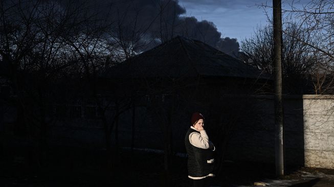 Seorang wanita berdiri di jalan saat asap hitam mengepul dari bandara militer di Chuguyev dekat Kharkiv pada 24 Februari 2022. Aris Messinis / AFP