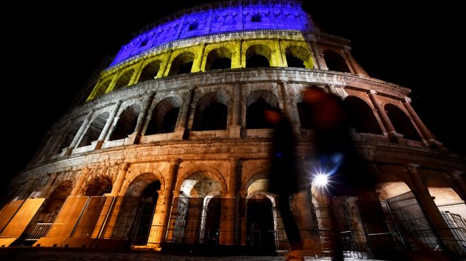 Pejalan kaki berjalan melewati bangunan Colosseum yang diterangi warna bendera Ukraina di Roma, Italia, Kamis (24/2/2022). [Filippo MONTEFORTE / AFP]