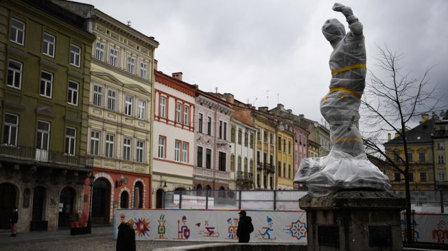 Warga berjalan melintasi patung bersejarah yang dibungkus di dekat gedung dewan kota di Lviv, Ukraina, Sabtu (5/3/2022). [Daniel LEAL / AFP]