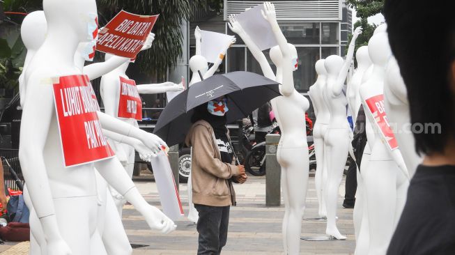 Seorang aktivis mengenakan masker bertanda silang berdiri disamping manekin saat melakukan aksi di depan Polda Metro Jaya, Jakarta Selatan, Senin (21/3/2022). [Suara.com/Alfian Winanro]