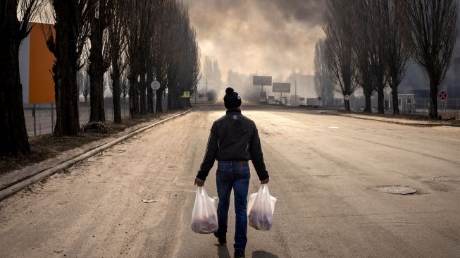 Seorang pria berjalan sambil memegang tas belanja di dekat gudang yang terbakar akibat dihantam rudal Rusia di pinggiran kota Kyiv, Ukraina, kamis (24/3/2022). [FADEL SENNA / AFP]