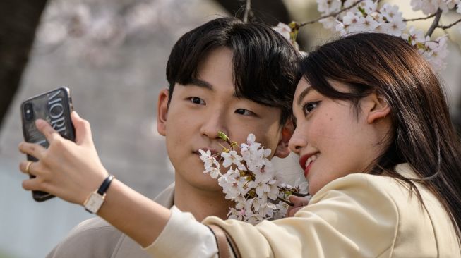 Sepasang kekasih berswafoto di samping pohon sakura yang bermekaran di Seoul, Korea Selatan, Rabu (6/4/2022). [ANTHONY WALLACE / AFP]