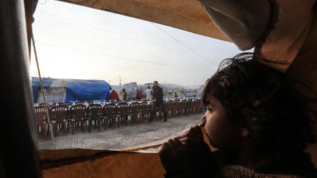 Seorang anak melihat dari dalam tenda ketika seorang sukarelawan menyiapkan meja bagi pengungsi Suriah untuk berbagi makanan berbuka puasa bersama saat bulan suci Ramadhan di Kota Al-Bab, provinsi Aleppo, Suriah, Kamis (7/4/2022). [Bakr ALKASEM / AFP]