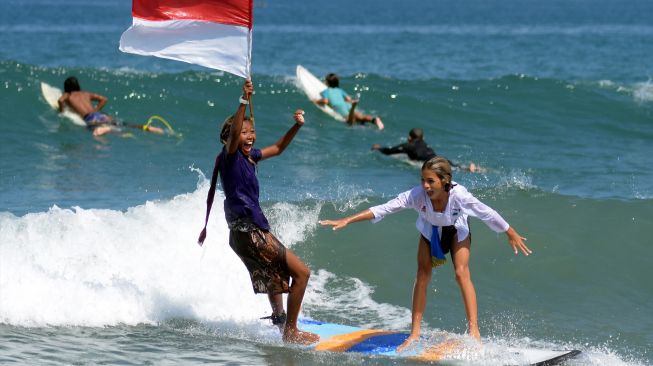 Dua peselancar wanita mengenakan pakaian kebaya sambil mengibarkan bendera Merah Putih saat beratraksi di Pantai Kuta, Badung, Bali, Sabtu (16/4/2022). [ANTARA FOTO/Nyoman Hendra Wibowo/rwa]
