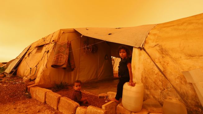 Anak-anak pengungsi duduk di dekat tenda mereka saat badai pasir melanda di atas kota Zardana, provinsi Idlib, Suriah, Kamis (2/6/2022). [Abdulaziz KETAZ / AFP]