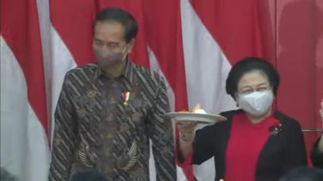 Presiden Joko Widodo atau Jokowi menyerahkan potongan tumpeng untuk Ketua Umum PDIP Megawati Soekarnoputri di acara Rakernas ke-2 PDIP di Sekolah Partai, Lenteng Agung, Jakarta Selatan, Selasa (21/6/2022). (Tangkap Layar YouTube).