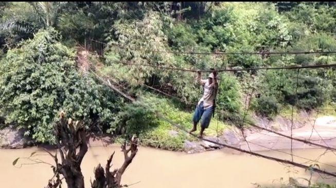 Seorang warga tengah melintasi jembatan rusak di atas Sungai Cisokan, Kabupaten Cianjur dan hanya berpijak pada seutas tali besi. [Suara.com/Fauzi Noviandi]