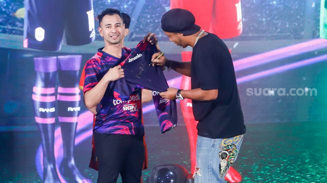 Legenda Sepak Bola Brazil, Ronaldinho (kanan) menandatangani Jersey baru musim 2022-2023 yang dipegang Chairman RANS Nusantara FC, Raffi Ahmad (kiri) di Jakarta, Jum'at (24/6/2022). [Suara.com/Alfian Winanto]