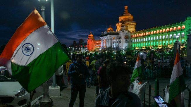 Seorang pedagang bendera mencari pelanggan di depan Vidhana Soudha, gedung legislatif negara bagian Karnataka yang diterangi warna bendera India di Bangalore, India, Sabtu (13/8/2022). [Manjunath Kiran / AFP]