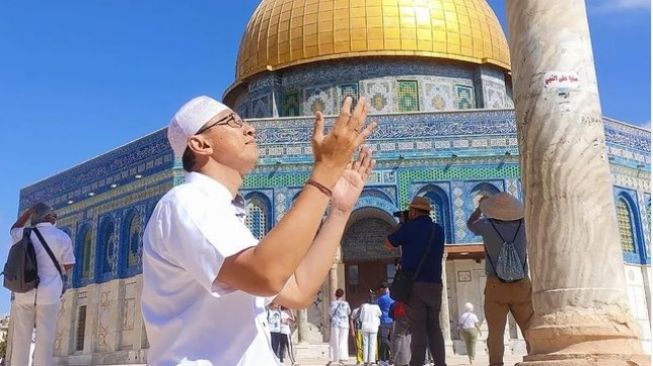 Di depan masjid Al-Aqsa, Abu Janda yang mengenakan pakaian serta peci putih terlihat sedang berpose orang yang sedang berdoa. [Instagram @permadiaktivis2]