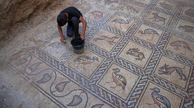 Putra petani Palestina Salman al-Nabahin saat membersihkan mozaik Bizantium yang ditemukan oleh ayahnya ketika menggali tanah di Bureij, Gaza, Palestina, Minggu (18/9/2022). [AFP PHOTO/MOHAMMED ABED]
