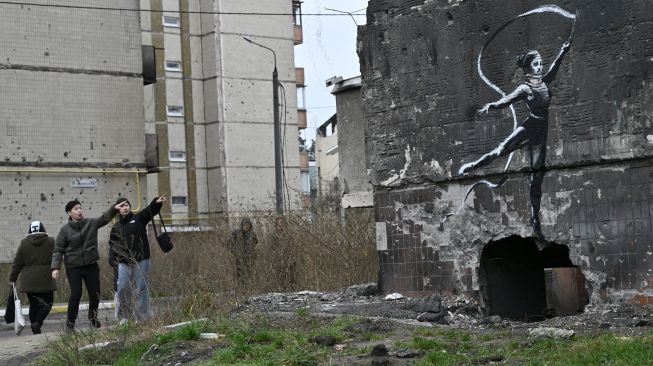 Penduduk setempat melihat grafiti Banksy di dinding bangunan tempat tinggal yang hancur, di Irpin, dekat Kyiv, Ukraina, Sabtu (12/11/2022). [Genya SAVILOV / AFP]