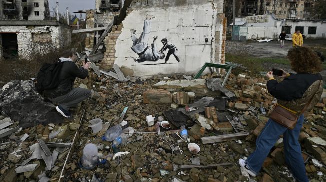 Orang-orang memotret grafiti Banksy di dinding sebuah bangunan yang hancur di Borodyanka, dekat Kyiv, Ukraina, Sabtu (12/11/2022). [Genya SAVILOV / AFP]