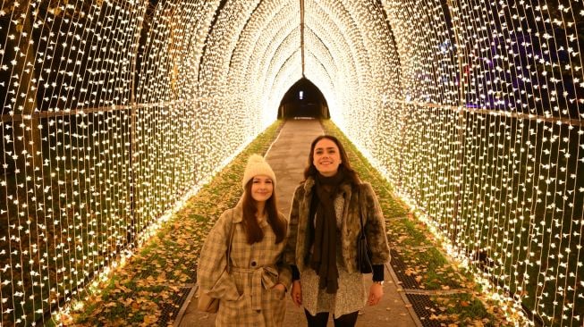 Pengunjung berpose di bawah instalasi cahaya saat perayaan natal Kew atau "Christmas at Kew" di Kew Gardens, barat daya London, Inggris, Selasa (15/11/2022). [Daniel LEAL / AFP]