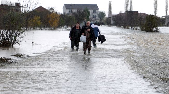 Orang-orang berjalan melalui jalan yang terendam banjir di desa Kuc dekat kota Shkodra, Albania, Senin (21/11/2022). [Gent SHKULLAKU / AFP]
