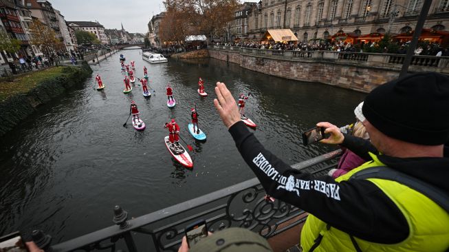 Seorang pejalan kaki mengambil gambar saat para pendayung berpakaian Sinterklas menghadiri parade SUP (Stand Up Paddleboarding) di sungai Ill di Strasbourg, Prancis, Sabtu (3/12/2022). [SEBASTIEN BOZON/AFP]