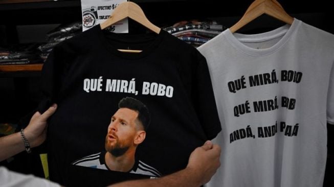 Seorang warga memegang kaus bergambarkan striker Argentina Lionel Messi dan frase yang berarti "Apa yang kau lihat, bodoh?" di suatu toko di Buenos Aires, Argentina. (12/12/2022) [AFP/LUIS ROBAYO]