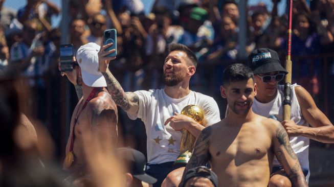 Bintang sepak bola Argentina Lionel Messi (tengah) berswafoto dengan ponselnya saat merayakan di atas bus dengan tanda bertuliskan "Juara Dunia" bersama pendukung setelah menjuarai turnamen Piala Dunia 2022 Qatar saat mereka melakukan tur keliling di pusat kota Buenos Aires, Argentina, Selasa (20/12/2022). [TOMAS CUESTA / AFP]