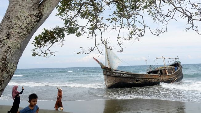 Warga menyaksikan kapal kayu menggunakan layar yang mana mengangkut puluhan imigran etnis Rohingya terdampar di dalam pantai Desa Ladong, Wilayah Aceh Besar, Aceh, Mingguan (25/12/2022). [ANTARA FOTO/Ampelsa].