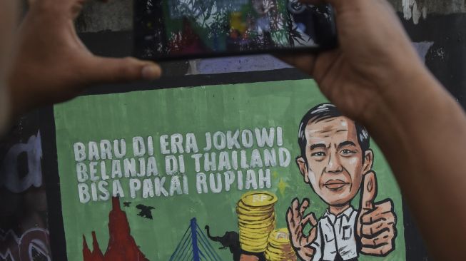 Warga memotret mural yang bertuliskan “Baru di era Jokowi belanja di Thailand bisa pakai rupiah” di Kabupaten Ciamis, Jawa Barat, Jumat (30/12/2022). [ANTARA FOTO/Adeng Bustomi].