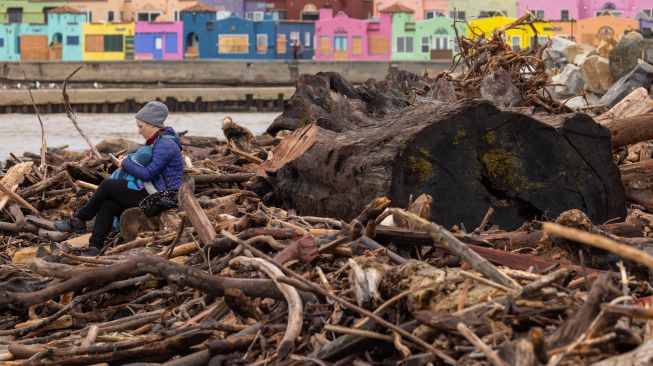 Seorang warga duduk bersama bayinya di antara pepohonan yang tersapu ke laut oleh badai dahsyat dan terdampar di pantai Capitola, California, Amerika Serikat Minggu (15/1/2023). [DAVID MCNEW / AFP]