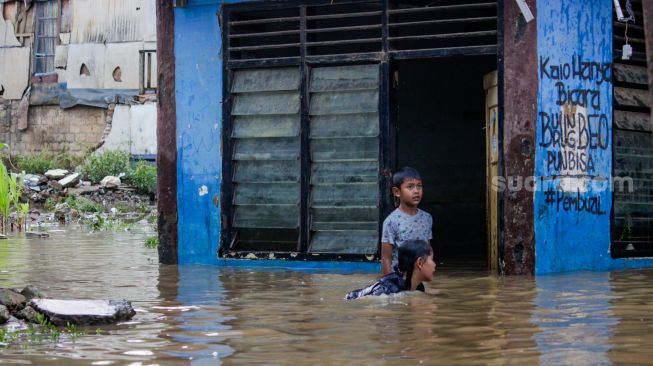 Anak-anak berjalan melewati banjir di kawasan pemukiman penduduk, Rawajati, Jakarta Selatan, Senin (27/2/2023). [Suara.com/Alfian Winanto]