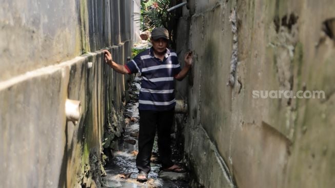 Ngadenin (63) berjalan melalui selokan untuk menuju rumahnya yang akses jalannya ditembok di kawasan Jatiwaringin, Bekasi, Jawa Barat, Senin (17/7/2023). [Suara.com/Alfian Winanto]