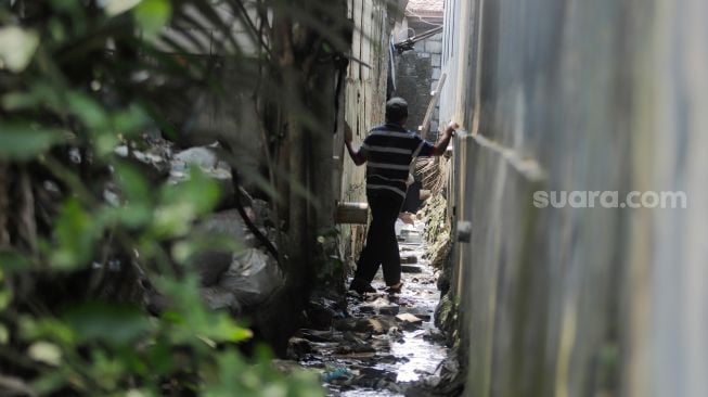 Ngadenin (63) berjalan melalui selokan untuk menuju rumahnya yang akses jalannya ditembok di kawasan Jatiwaringin, Bekasi, Jawa Barat, Senin (17/7/2023). [Suara.com/Alfian Winanto]