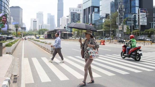 Pekerja melindungi tubuh dari terik matahari menggunakan payung saat berjalan di kawasan Sudirman, Jakarta, Kamis (21/12/2023). [Suara.com/Alfian Winanto]