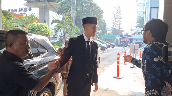 Jay Idzes saat tiba di Kanwil Kemenkumham Jakarta didampingi oleh wanita berambut pirang. Nampak Jay mengenakan setelan jas hitam, kemeja putih, dan pakai peci untuk menjalani sumpah WNI, Kamis (18/12/2023). (Suara.com/Adie Prasetyo Nugraha).