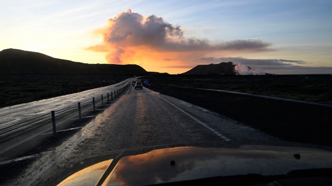 Penampakan asap letusan gunung berapi yang terlihat dibalik lanskap jalan yang menghubungkan Reykjavik dan Grindavik di Islandia barat, Minggu (14/1/2024). [Halldor KOLBEINS / AFP]
