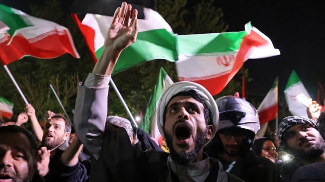 Demonstran mengibarkan bendera Iran dan bendera Palestina saat mereka berkumpul setelah Iran melancarkan serangan drone dan rudal ke Israel di depan Kedutaan Besar Inggris di Teheran, Iran, Minggu (14/4/2024). [ATTA KENARE / AFP]