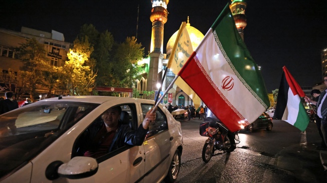 Demonstran mengibarkan bendera Iran dan bendera Palestina saat mereka berkumpul setelah Iran melancarkan serangan drone dan rudal ke Israel di Lapangan Palestina di Teheran, Iran, Minggu (14/4/2024). [ATTA KENARE / AFP]