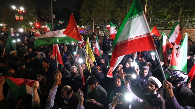 Demonstran mengibarkan bendera Iran dan bendera Palestina saat mereka berkumpul setelah Iran melancarkan serangan drone dan rudal ke Israel di depan Kedutaan Besar Inggris di Teheran, Iran, Minggu (14/4/2024). [ATTA KENARE / AFP]