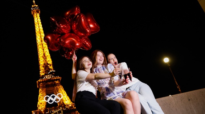 Wisatawan memegang balon berbentuk hati berwarna merah dan gelas anggur saat mereka berpose di depan Menara Eiffel yang dihiasi cincin Olimpiade di Paris, Prancis Jumat (7/6/2024). [Ludovic MARIN / AFP] 